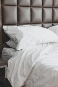 A photograph of the corner of a bed with white sheets and a grey, padded headboard.