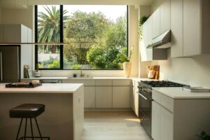 A photograph of a neutral color kitchen with a large island and big windows overlooking trees.